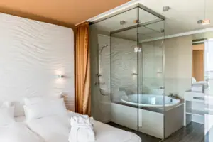 Suite at the aja Warnemünde A bathroom with a glass shower, bathtub and glass wall, with the bed in front of it.