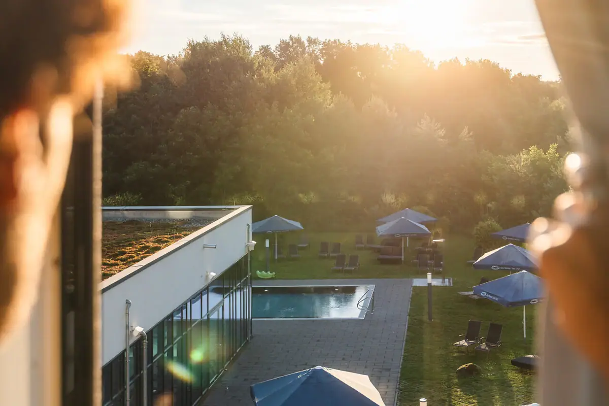 View from the room at aja Bad Saarow A person looks out of a window onto a pool with parasols.