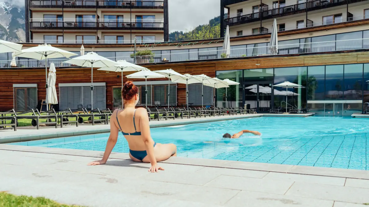A woman is sitting in a swimming pool whilst a man is swimming in it.
