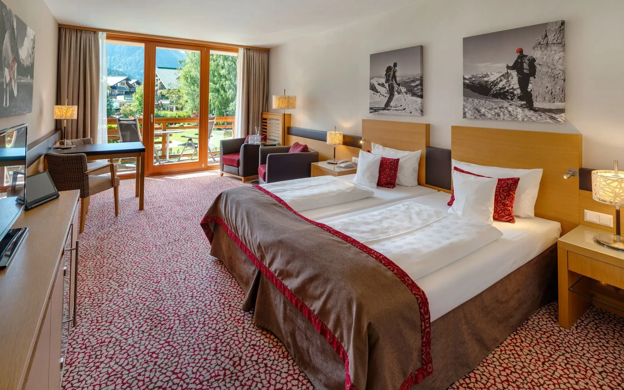 Hotel room with a bed, armchairs, desk and a balcony with a view of the mountains.