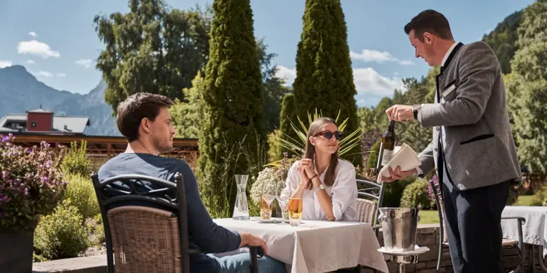 A man pours wine into a bottle at an outdoor table where people are sitting.