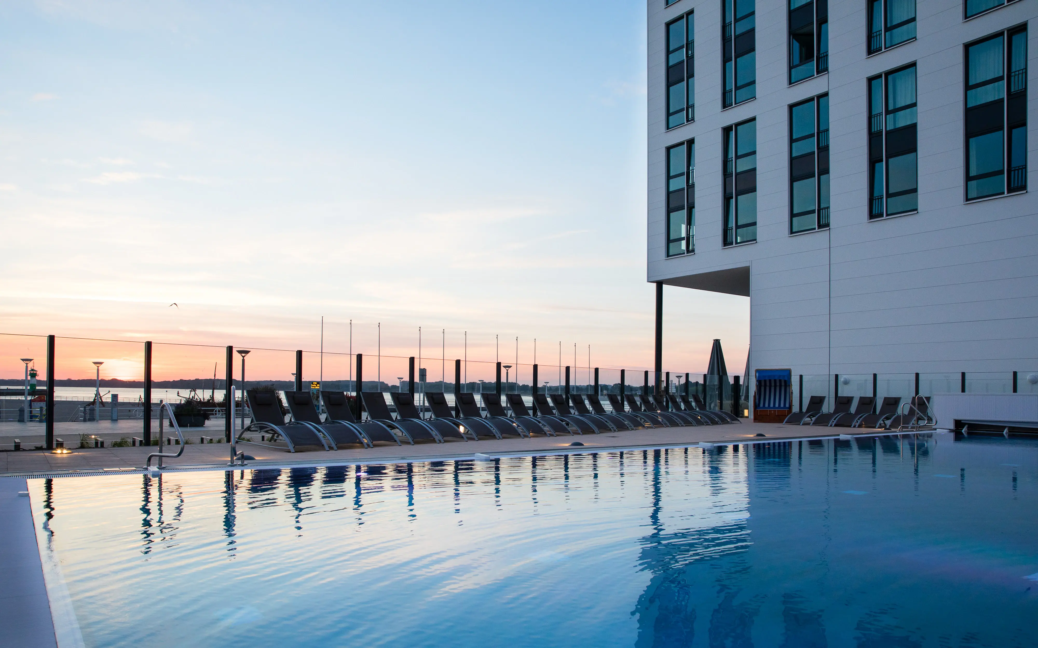 Outdoor pool area at sunset with rows of sun loungers and a modern hotel building, reflecting on the calm water.