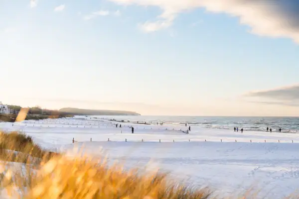 The beach in Warnemünde is covered in snow. The dunes glow brightly in the sun and people walk along the beach in warm clothes. 