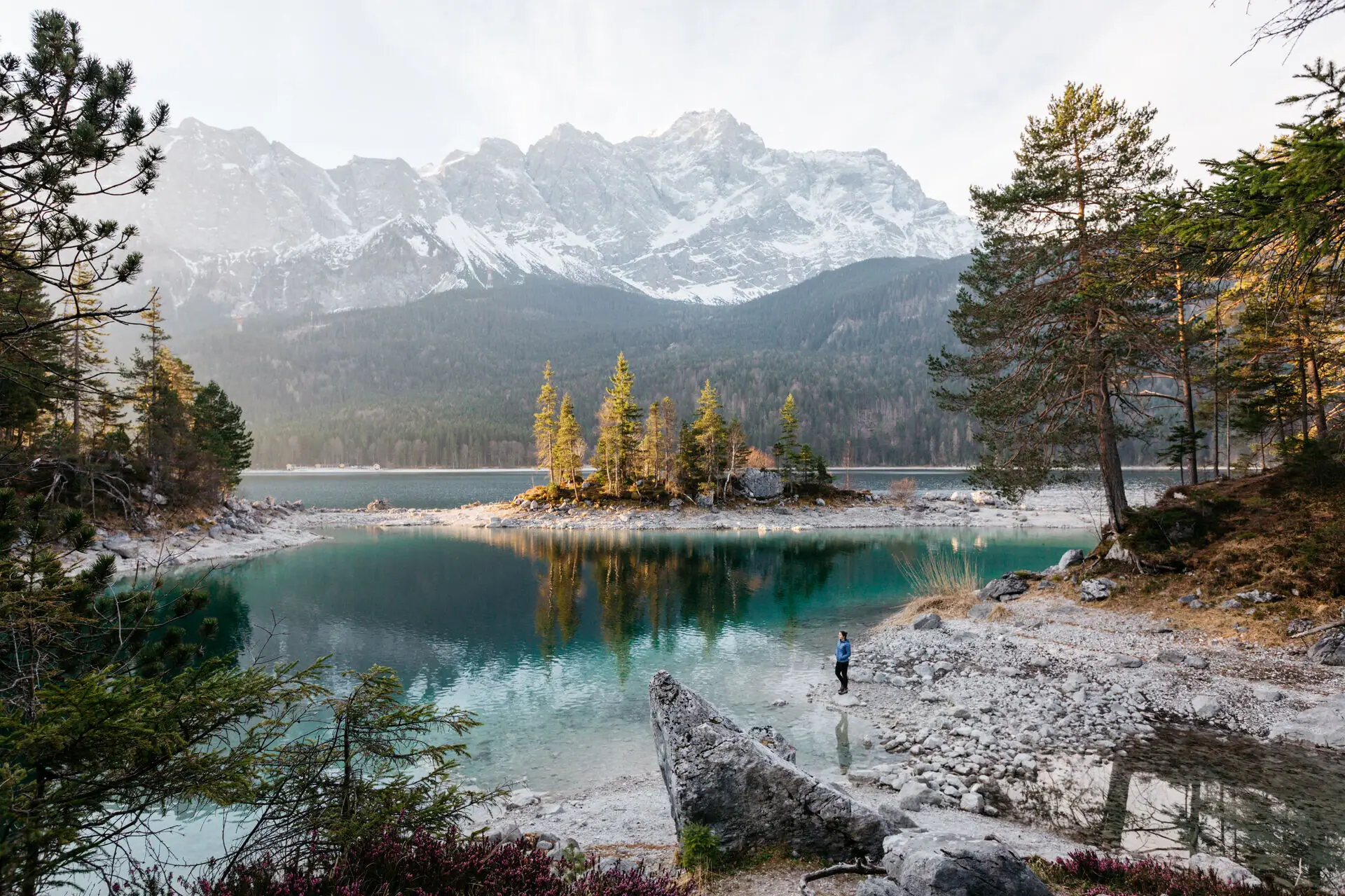 A person stands on a rocky shore with a small island in the centre of a lake.