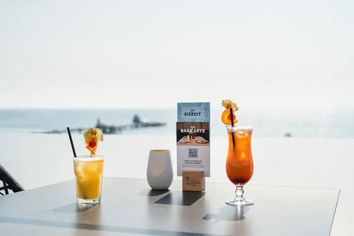 Two cocktails on a table in the aja Rooftop Bar Grömitz with a sea view and the pier in the background.