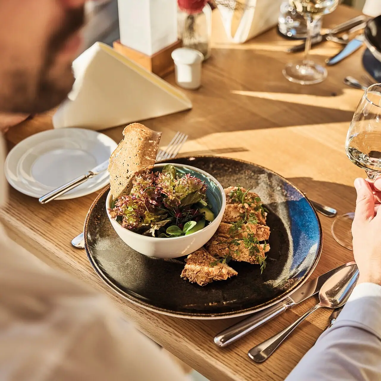 A man eats from a plate and holds a glass of wine. There are two dishes on the table. The sun is shining on the table.