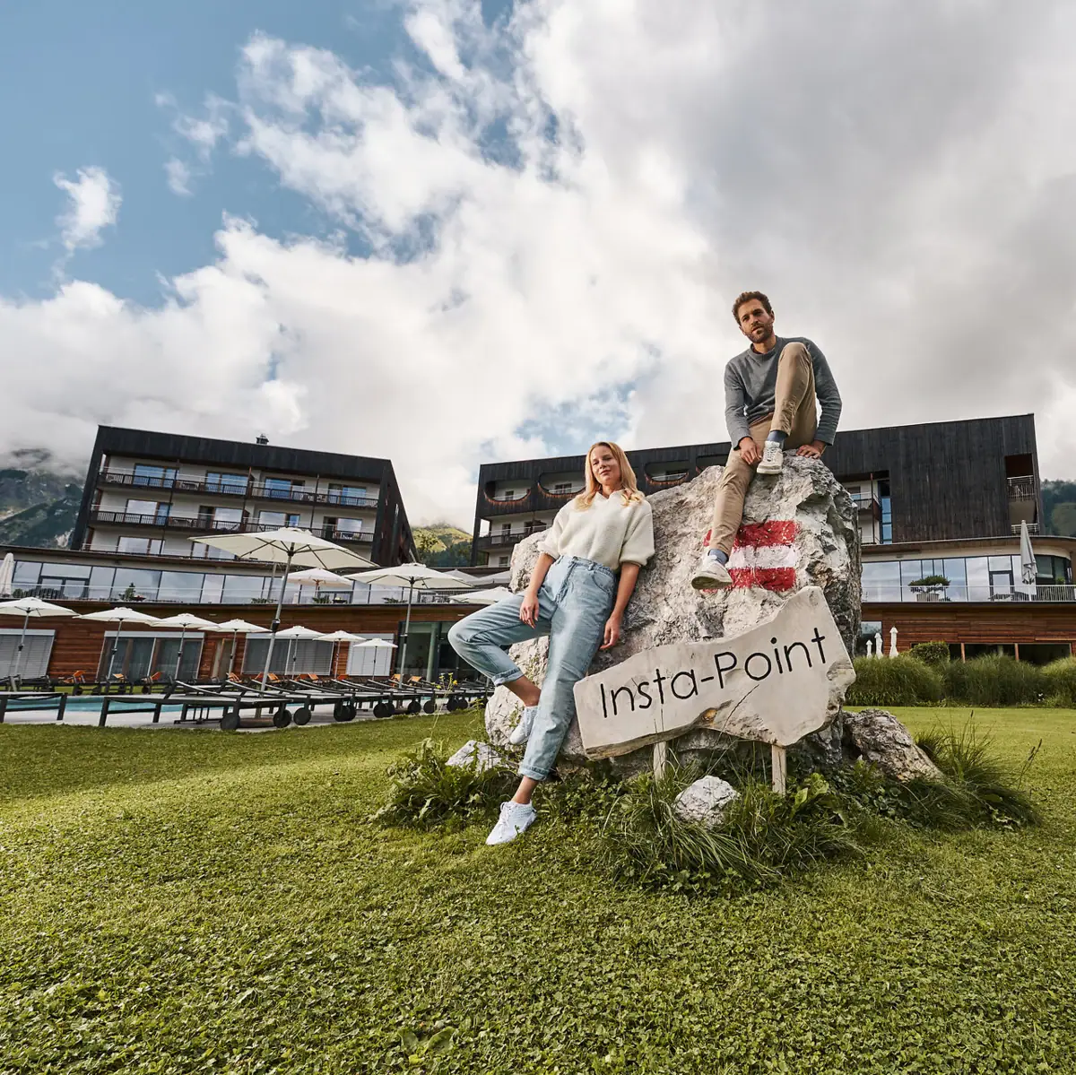 A man and a woman sit on a rock with a sign, the hotel in the background.