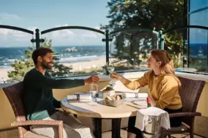 Ein Mann und eine Frau sitzen an einem Tisch mit Weingläsern mim Freien und im Hintergrund ist das Meer zu sehen..