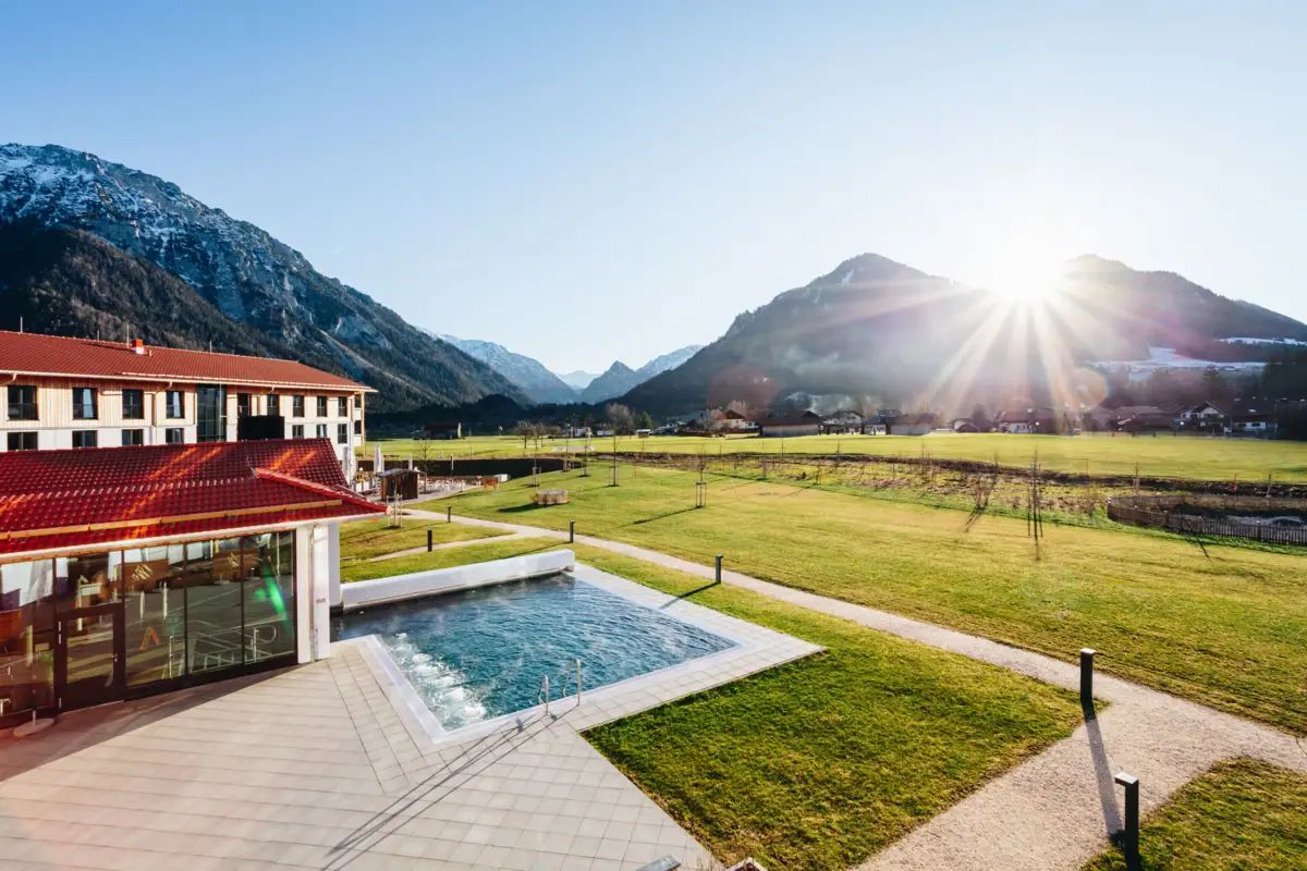 Ruhpolding Outdoor Pool The aja Ruhpolding, a wellness hotel in Bavaria with a swimming pool set in grassy surroundings against a backdrop of mountains.