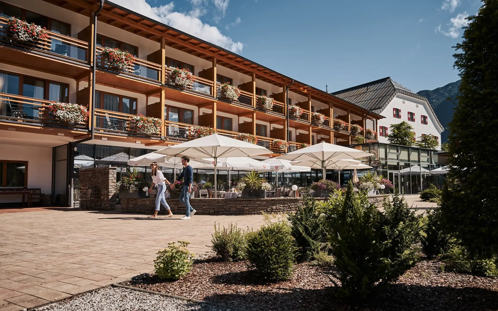 A couple strolls in front of a building with an outdoor terrace and large parasols.