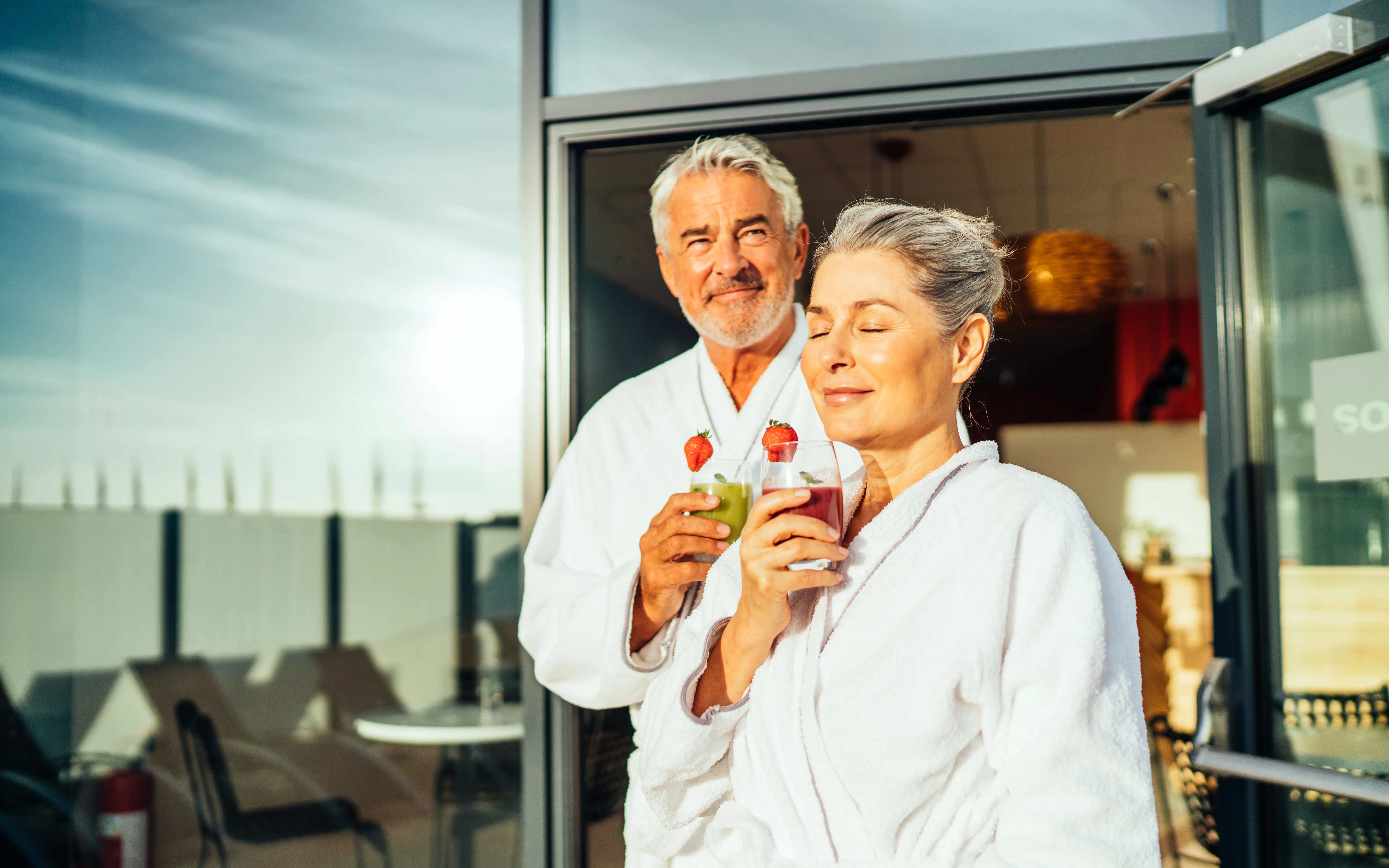 Elderly couple in bathrobes enjoying fresh fruit smoothies on a sunny terrace outside a wellness area.