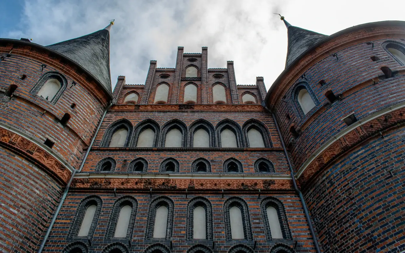 Close-up view of a historic brick building with intricate Gothic-style windows and two rounded towers, set against a cloudy sky.