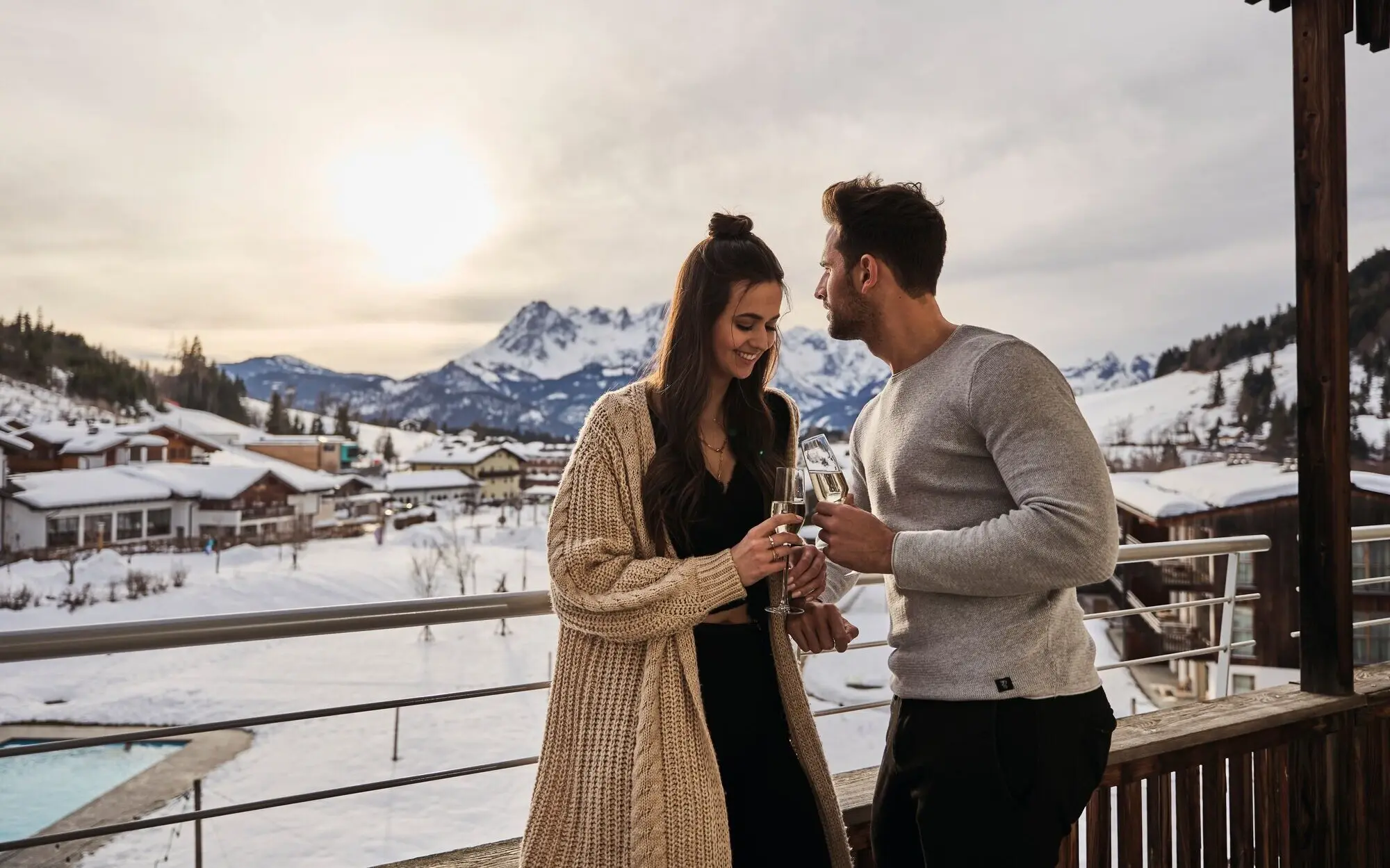 A man and a woman hold champagne glasses on a balcony with snow-covered mountains in the background.