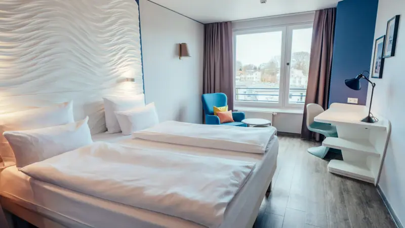 A room with two beds and a table, featuring neatly arranged bedding and pillows.