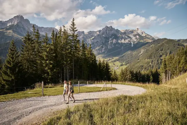 Two people are walking on a gravel path in front of a mountain range.