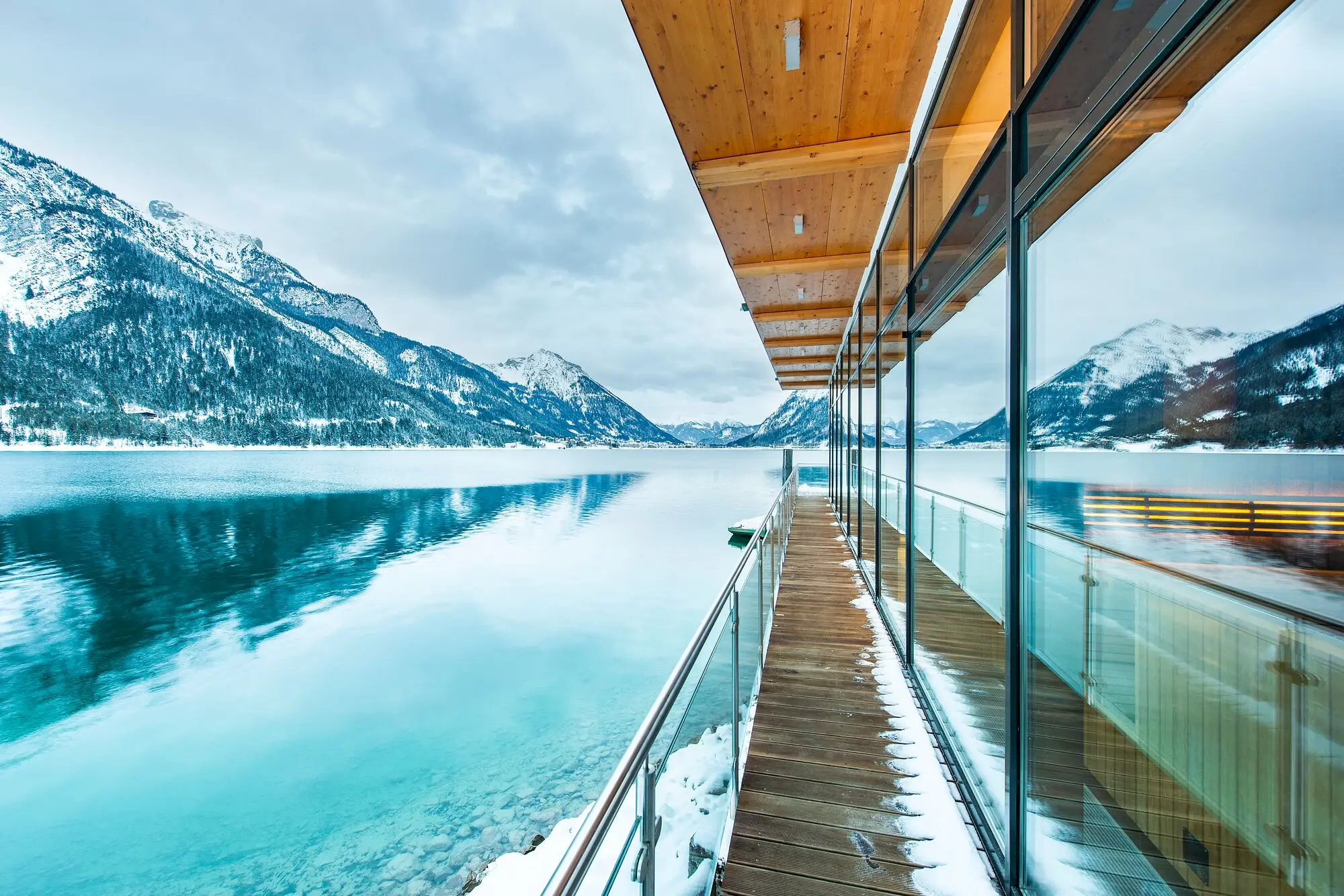 Glass wall with wooden deck overlooking a lake, surrounded by mountains and clouds.