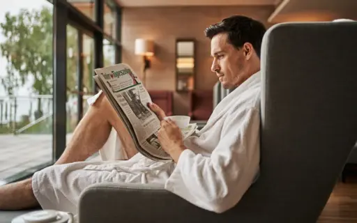 Man in a bathrobe reading a newspaper on a sofa.