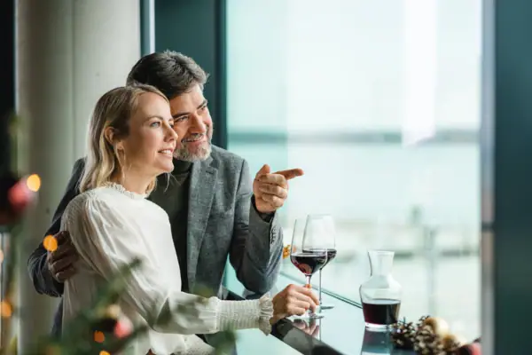 A man and a woman sit at a bar with glasses of wine. Christmas lights shine in the foreground.