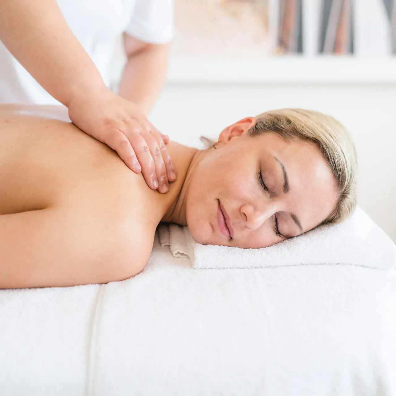 A woman lies on a white towel with her eyes closed and her neck is massaged.