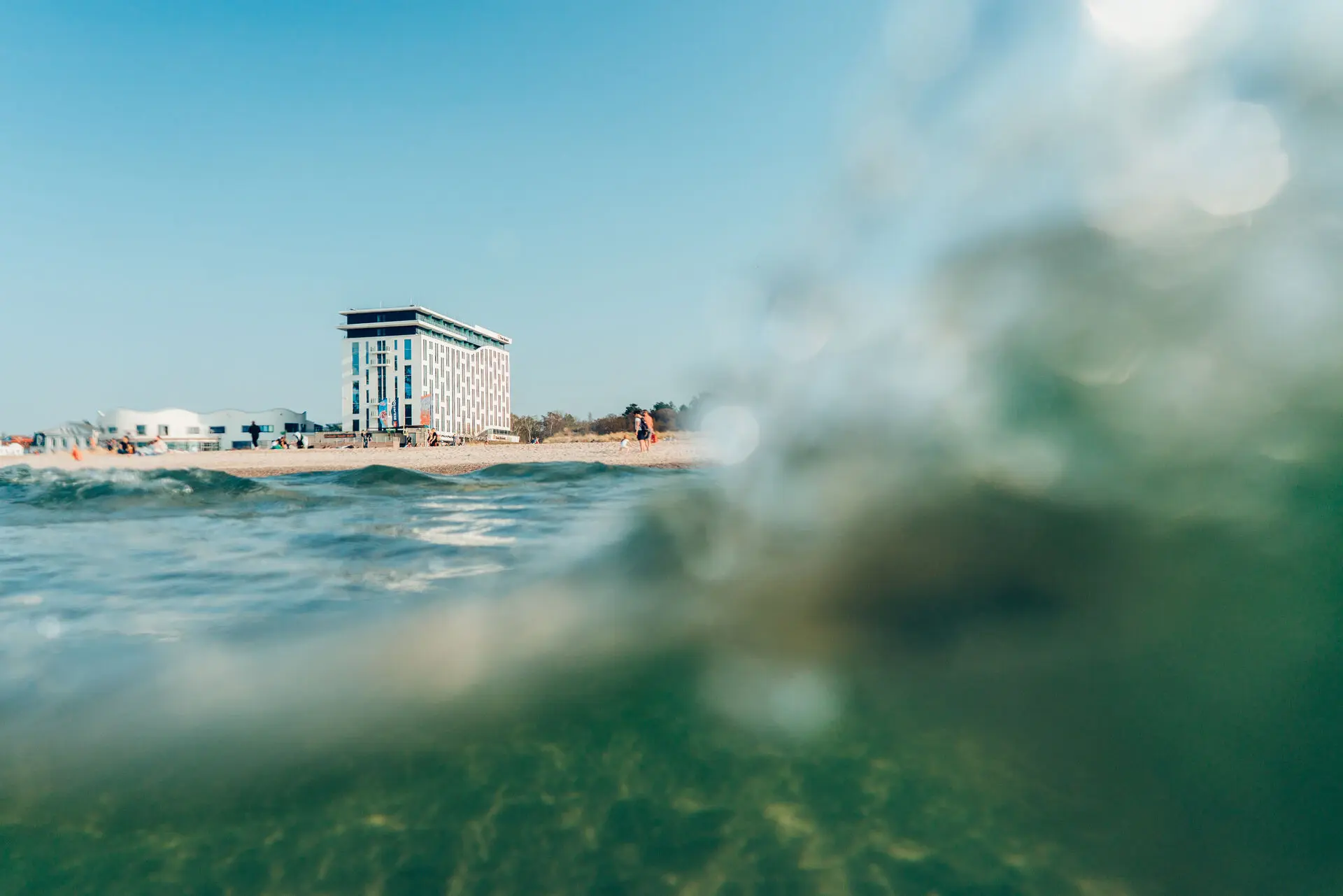 View from the water towards a beachfront hotel, with gentle waves in the foreground and a clear blue sky above, capturing a serene coastal atmosphere.