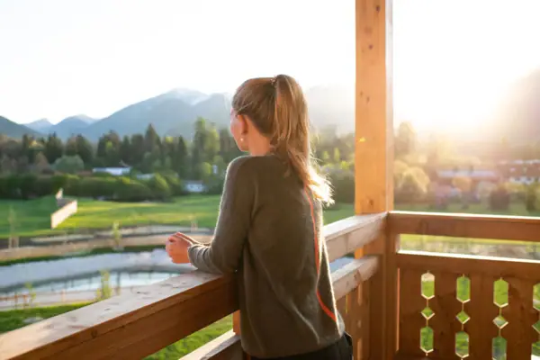 A woman leans on the wooden railing of a balcony and looks into the distance. The morning sun shines warmly into the picture. 
