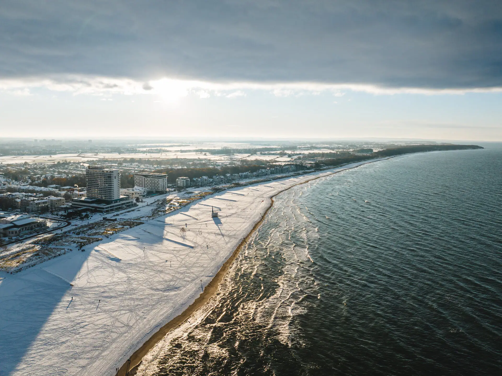 aja Warnemünde Ein schneebedeckter Strand mit Gebäuden und Wasser im Hintergrund.
