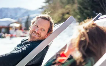 Man sitting on a deck chair outdoors, smiling and a snowy landscape can be seen in the background.