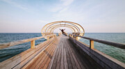 Person stands on a wooden footbridge by the water.