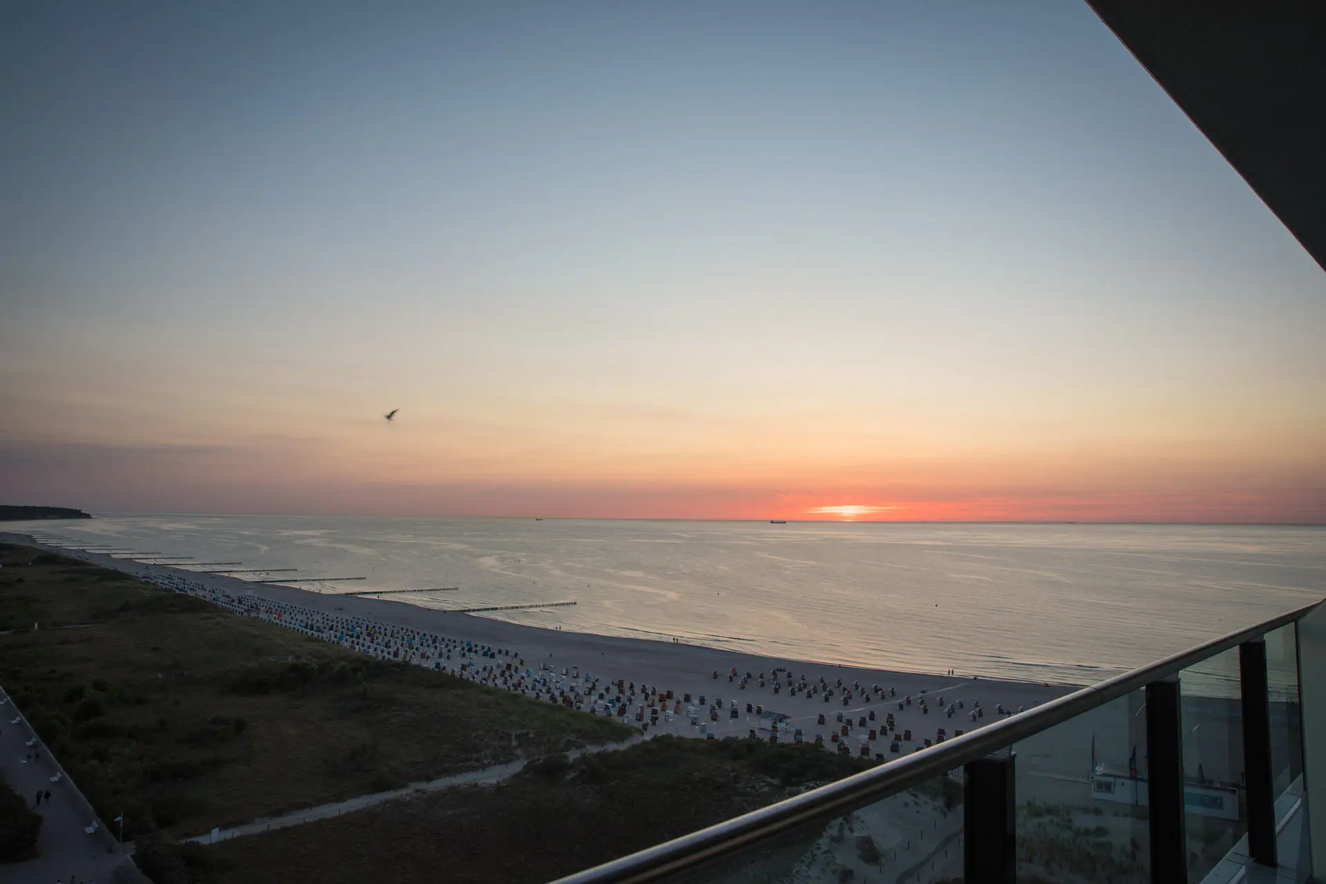 Sunset view from a balcony overlooking a sandy beach with beach chairs, calm sea waves, and a serene horizon, creating a tranquil coastal atmosphere.