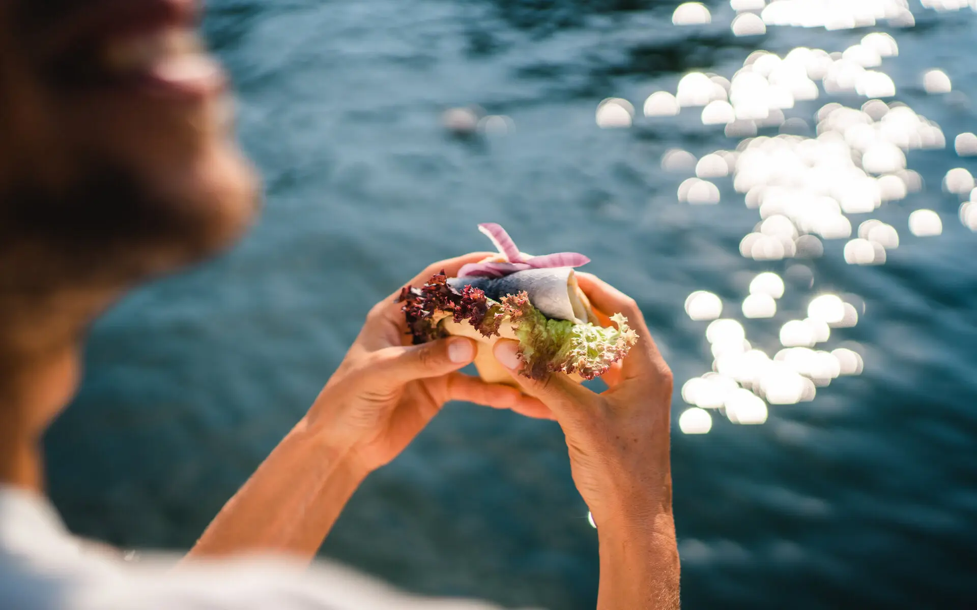 A woman ist standing by the water holding a fish sandwich in her hands. 