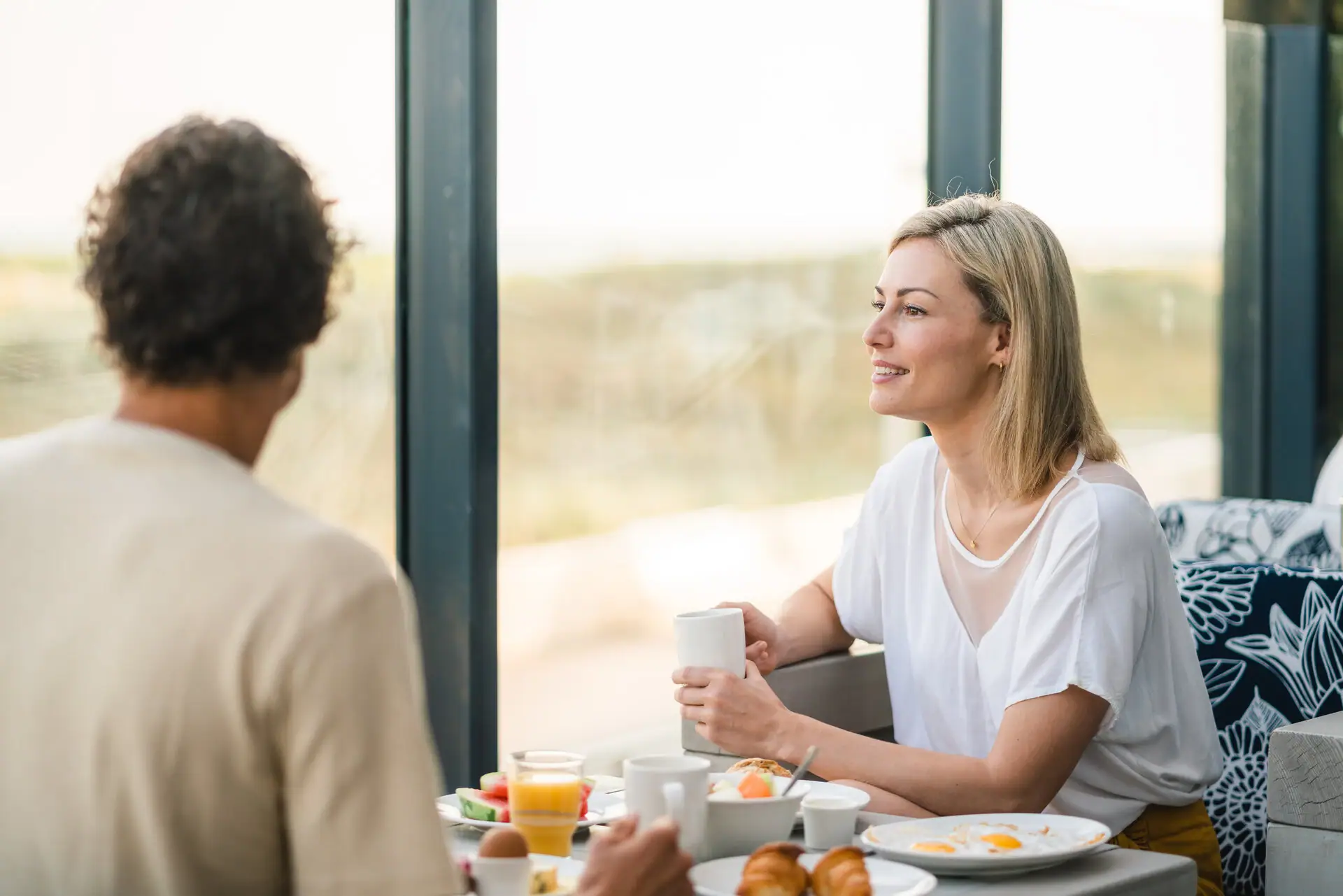 A couple is sitting at the breakfast table and one woman is holding a cup and looking at the other person. The dunes can be seen in the background.