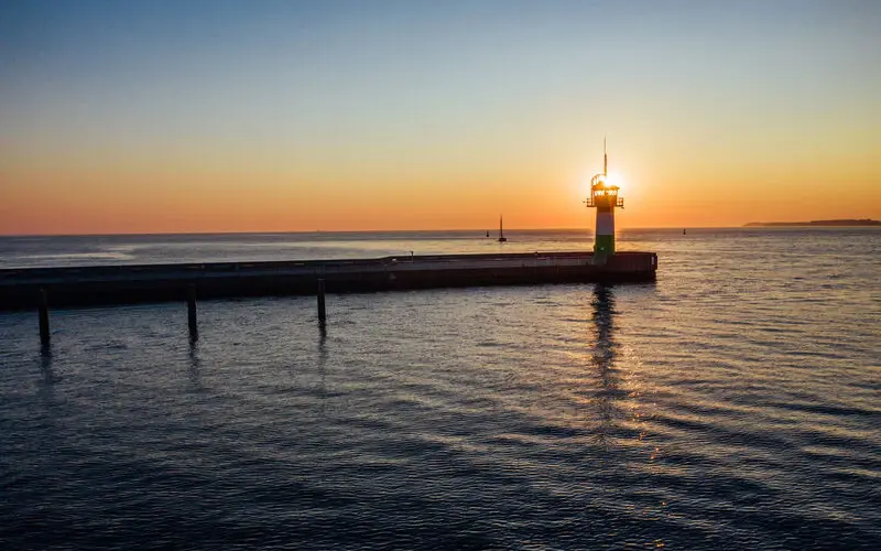 Scenic view of a green lighthouse on a pier at sunset, with the sun glowing behind it and calm ocean waters reflecting the warm hues of the sky.