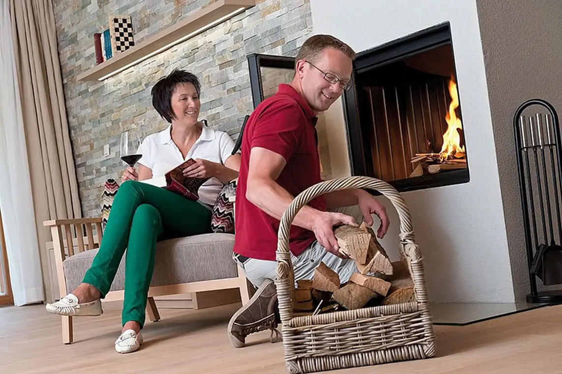 A man and a woman are sitting in front of a fireplace, the man is adding wood from a basket and the woman is holding a glass of red wine.