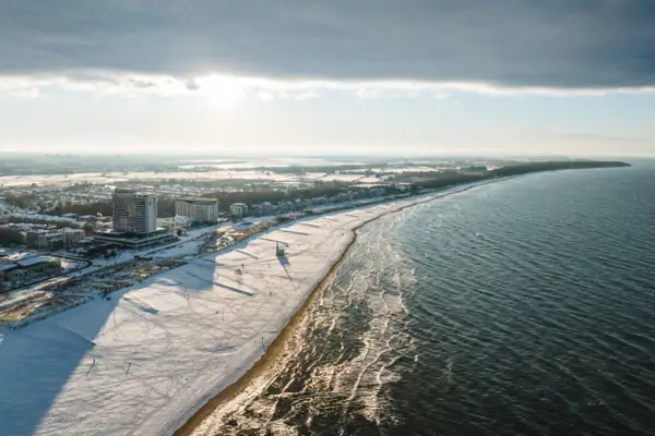 aja Warnemünde Ein schneebedeckter Strand mit Gebäuden und Wasser im Hintergrund.