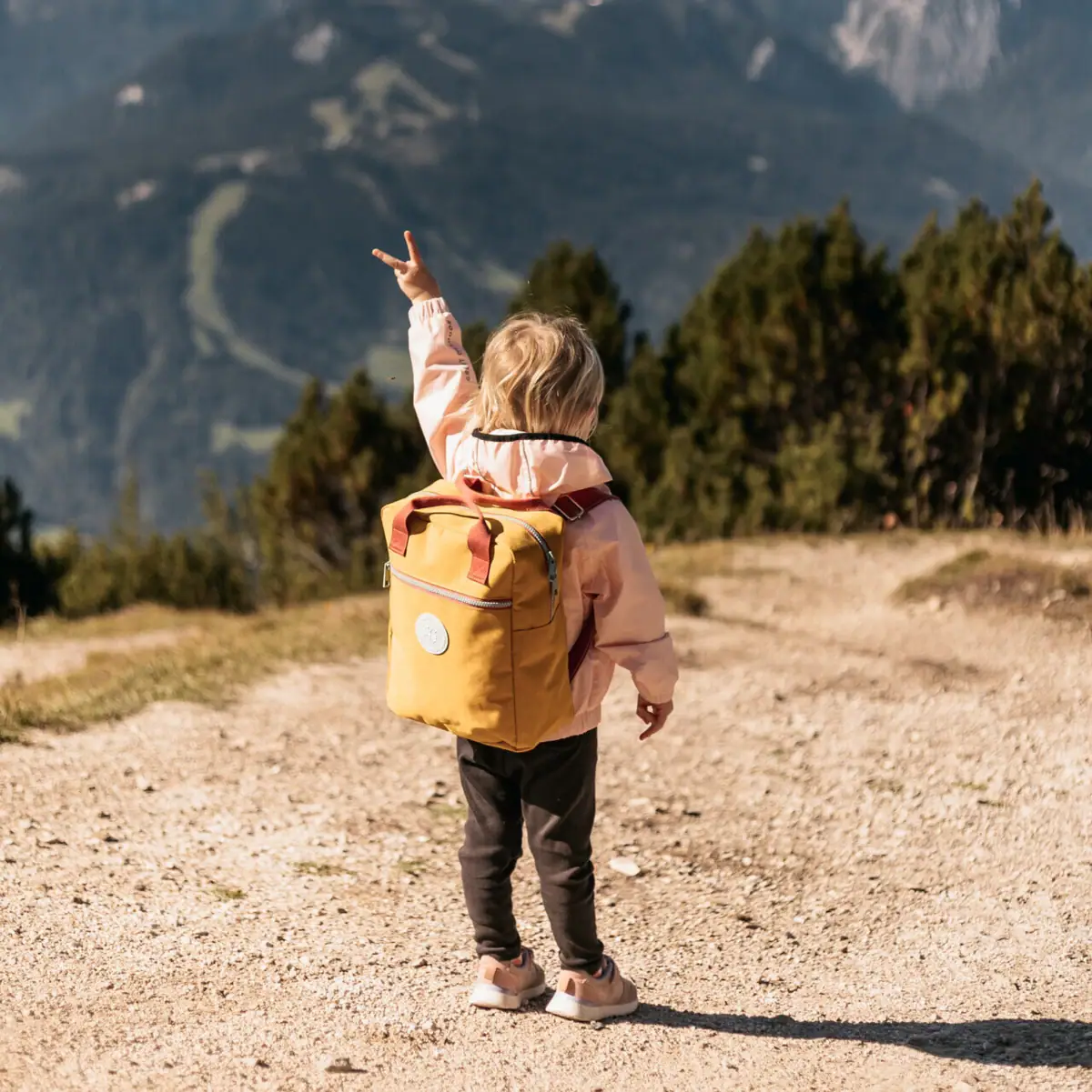 A child with a yellow rucksack stands on a dirt track, with a mountain range in the background.