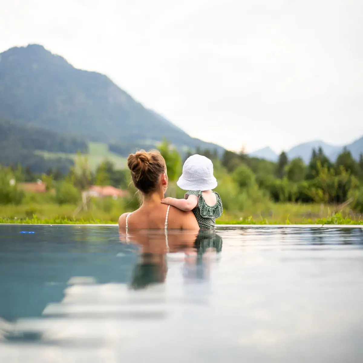 A mother sits with her toddler in the water of an infinity pool and looks out over an idyllic mountain landscape.