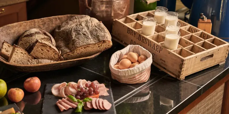 A breakfast buffet with a large bread basket, a platter, breakfast eggs, apples and glasses of milk.