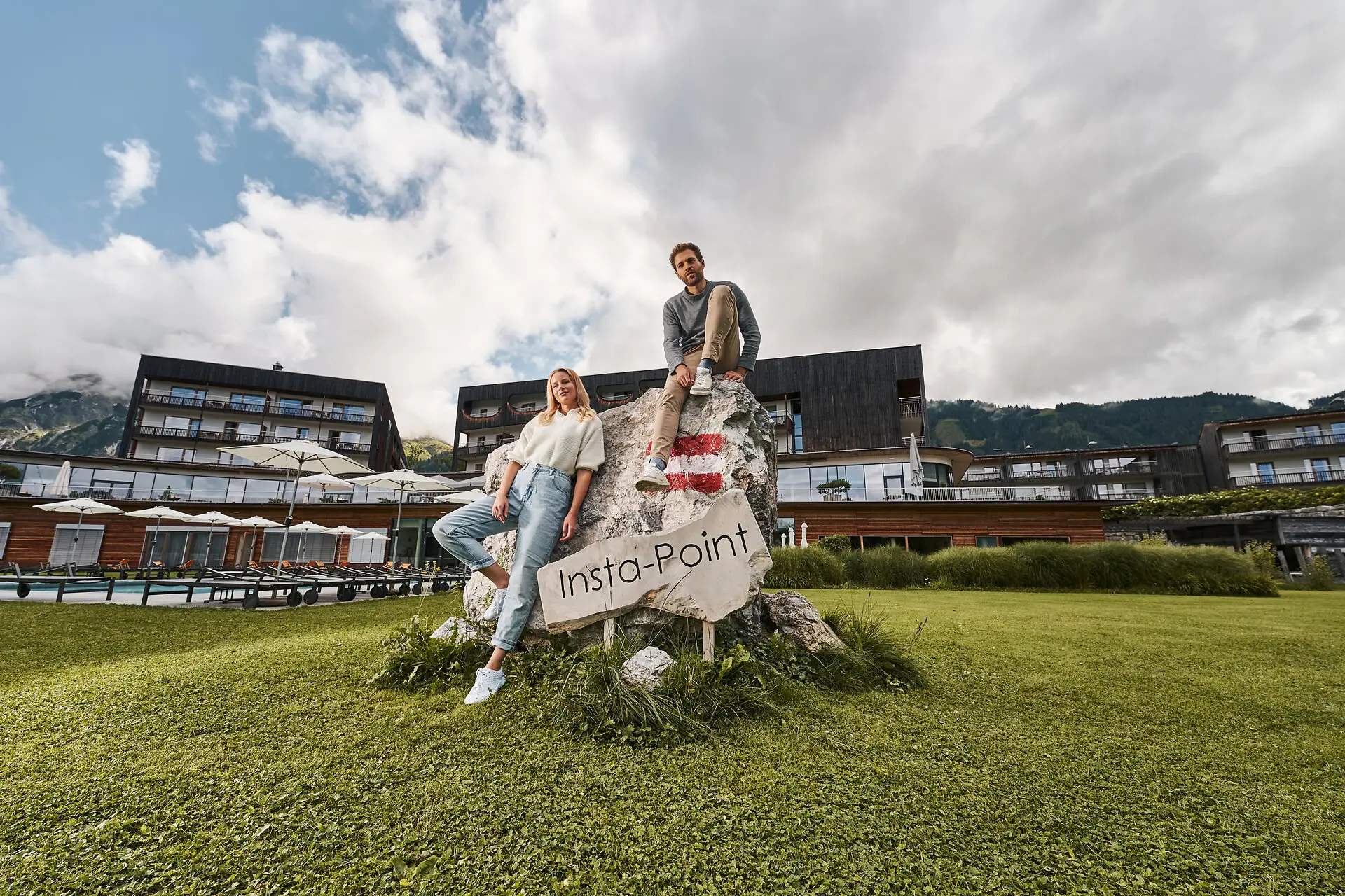 Ein Mann und eine Frau sitzen auf einem Felsen mit einem Schild, im Hintergrund das Hotelgebäude.