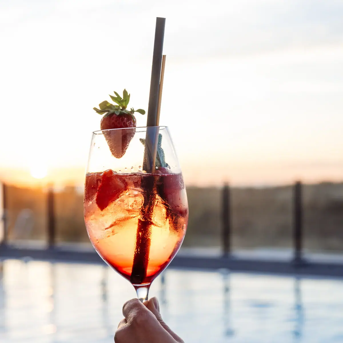 Drink One hand holds a drink with strawberries, a pool can be seen in the background.