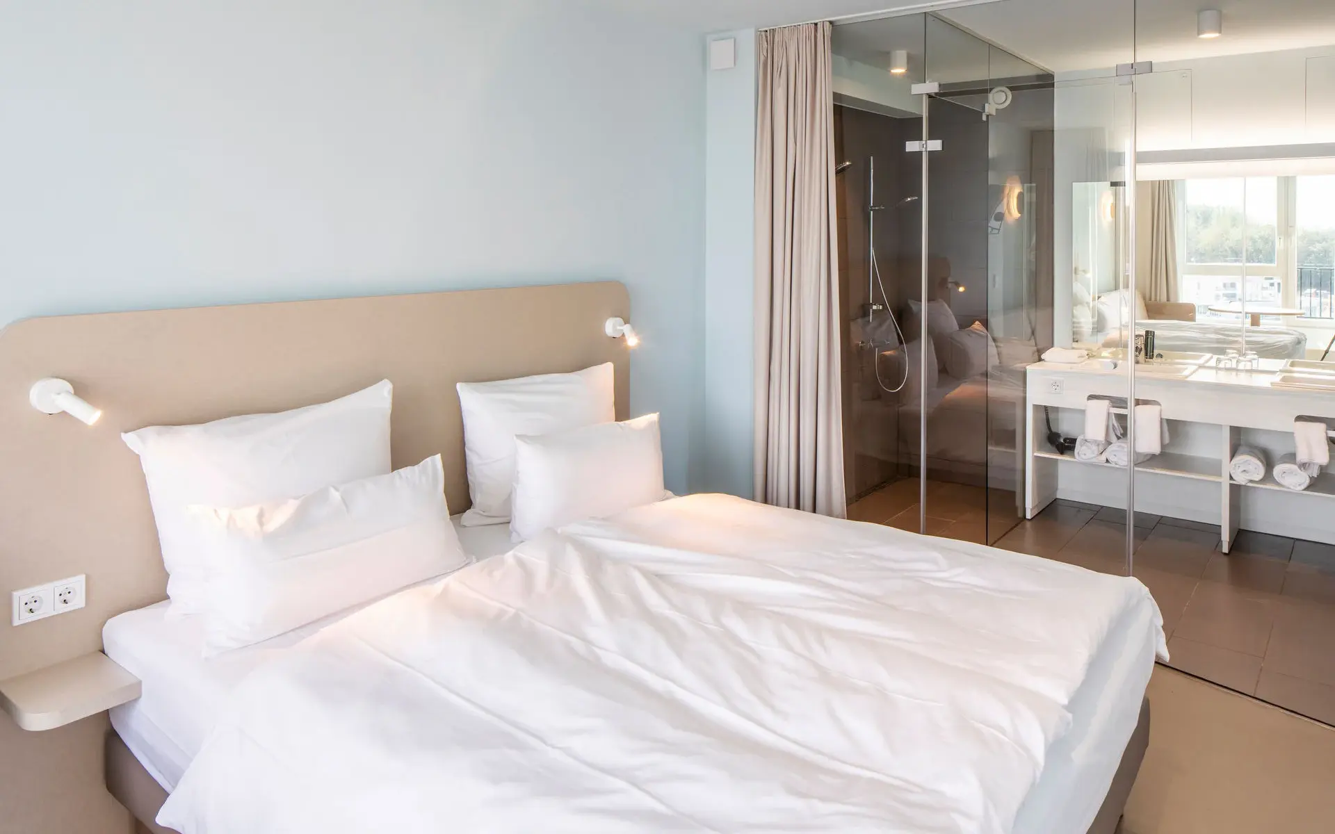 A bright hotel room with a large bed and a bathroom with a glass wall in the background. 