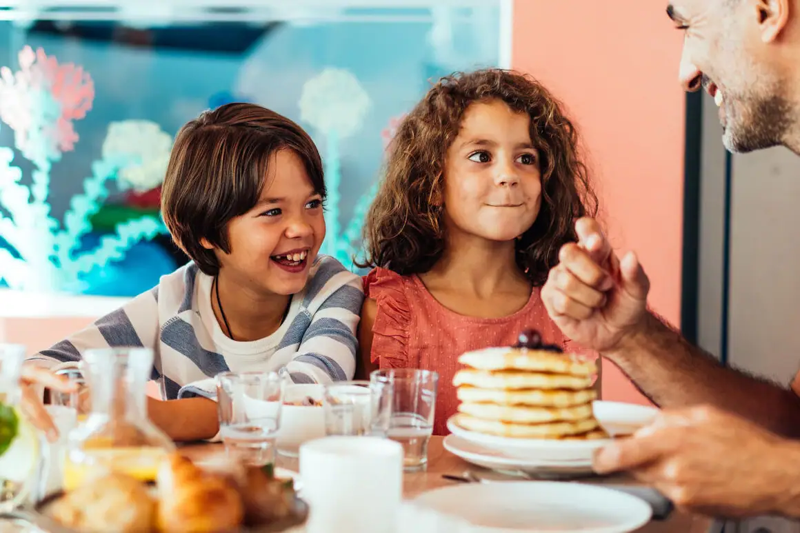Two children sit with their father in the family area at a table with pancakes, drinks and bread rolls.