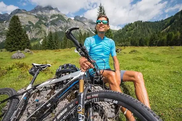 Man sitting on a rock with his bicycle in front of a mountain backdrop.