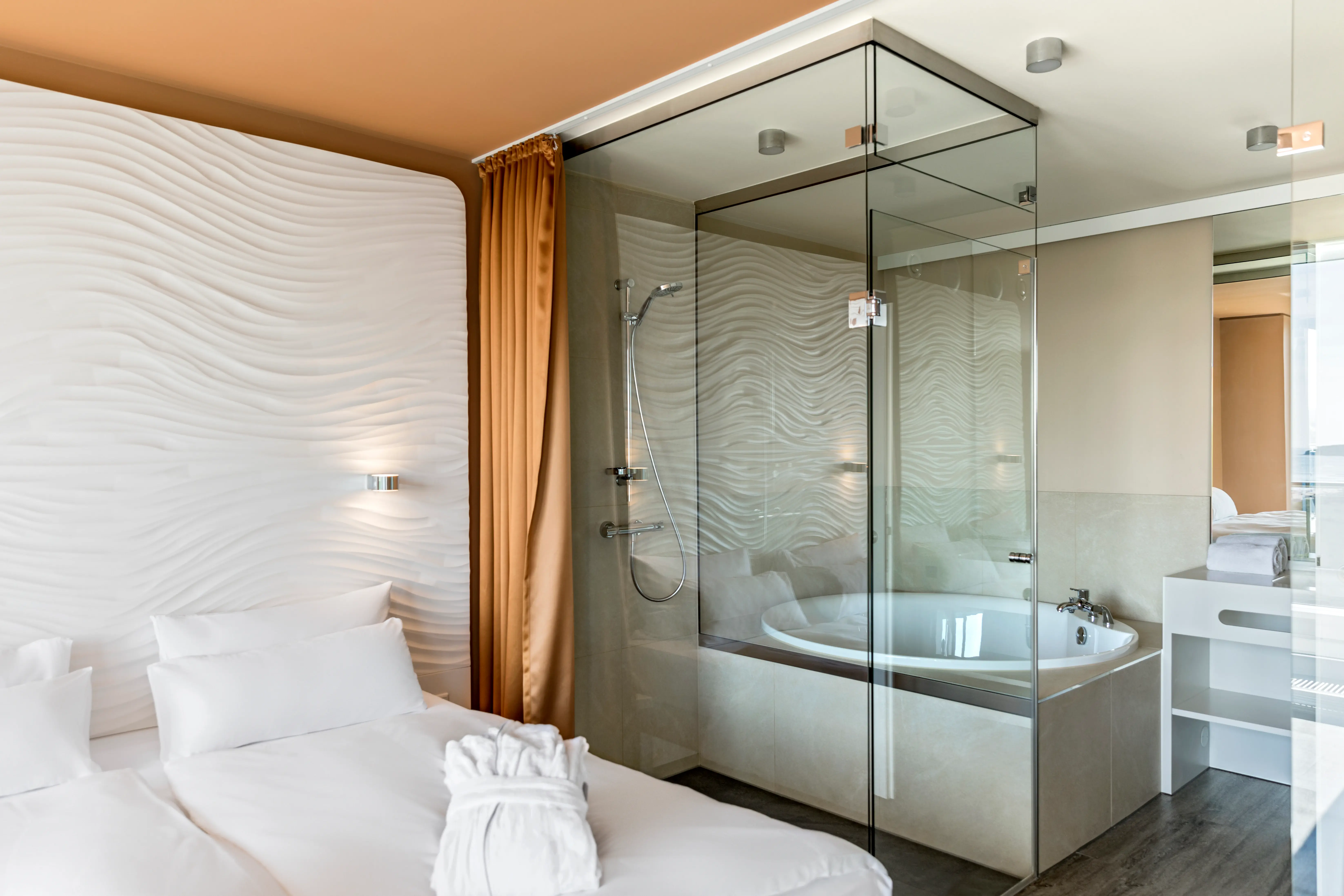 Modern hotel room with a comfortable double bed, stylish wave-patterned wall design, and an open-plan bathroom with glass partitions and a bathtub, creating a bright and luxurious atmosphere.