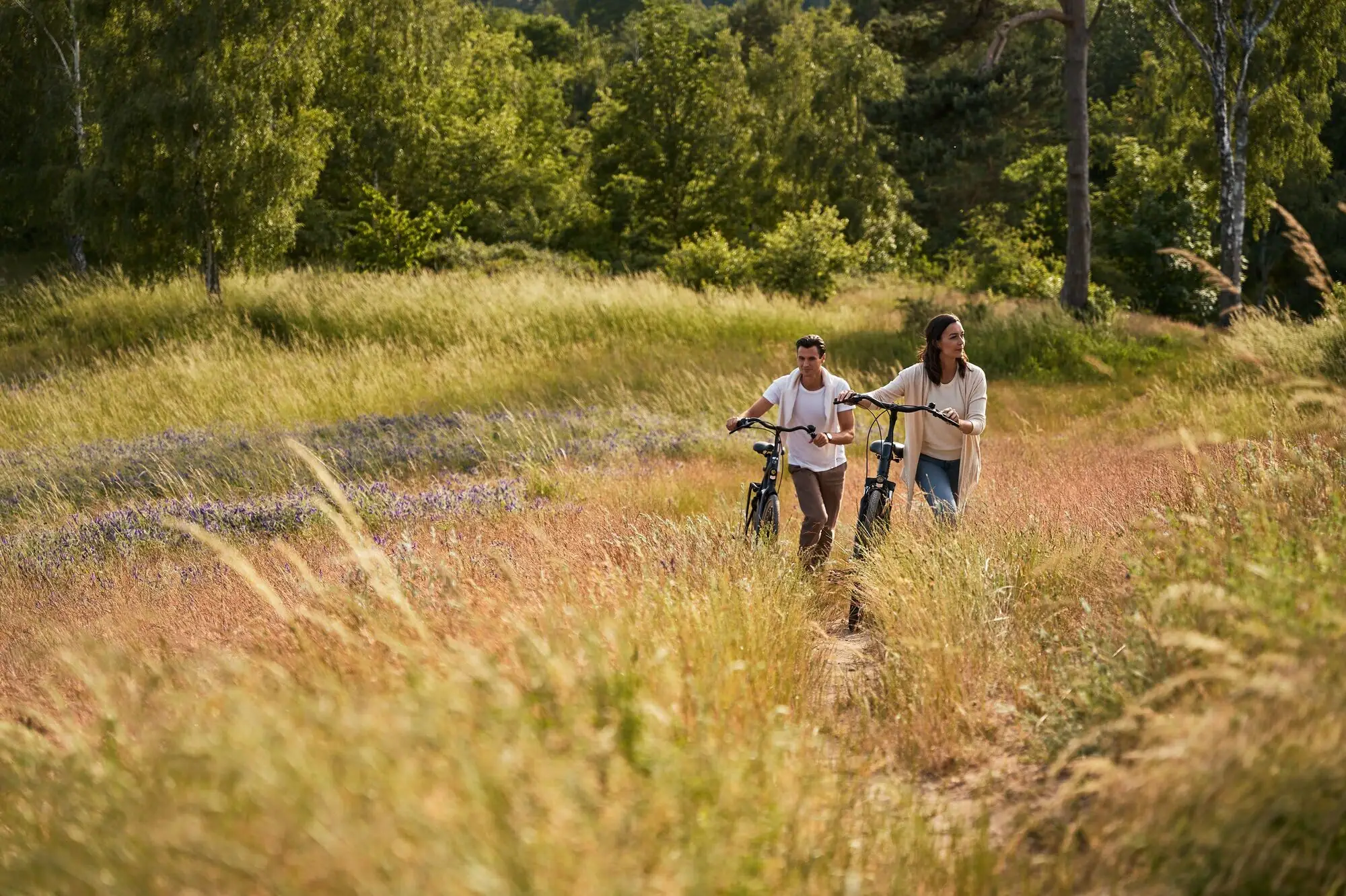A man and a woman are walking through a field on bicycles.