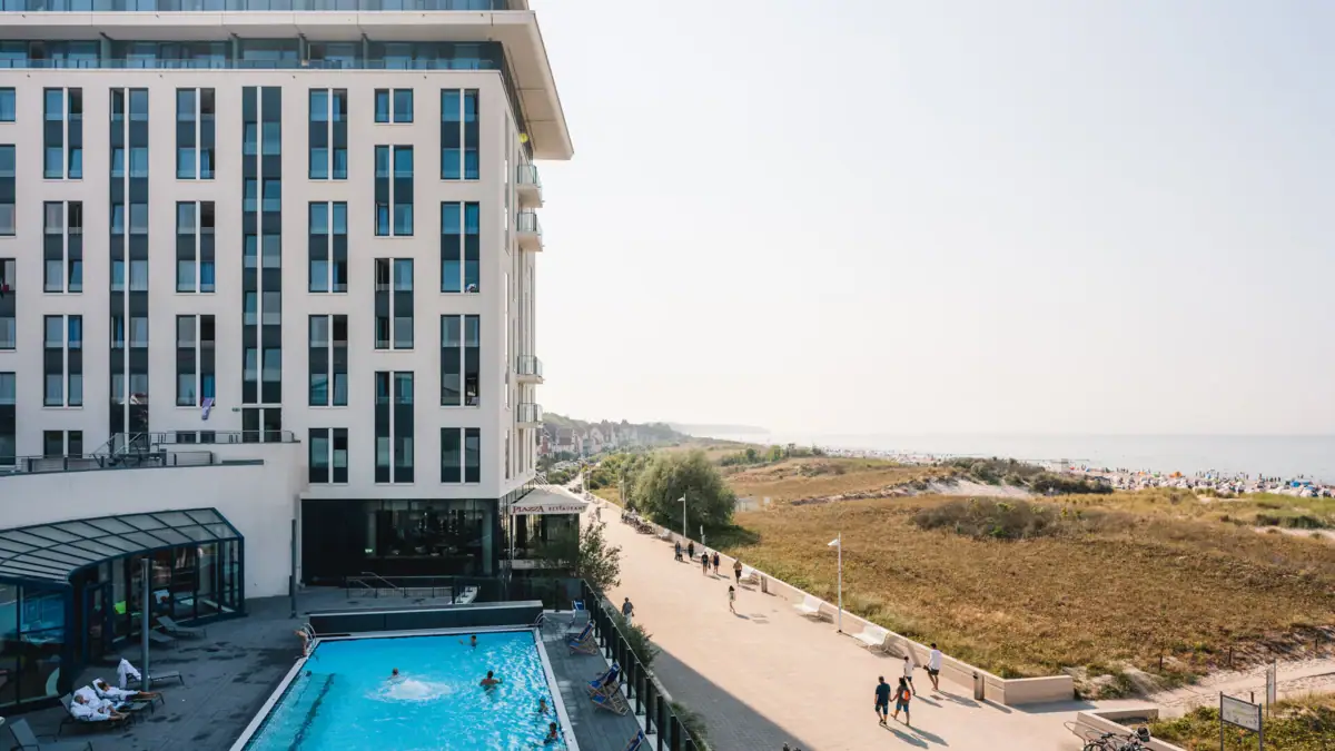 Wellness hotel aja Warnemünde A building with a pool, with people walking past. The sea and the beach can be seen in the background.