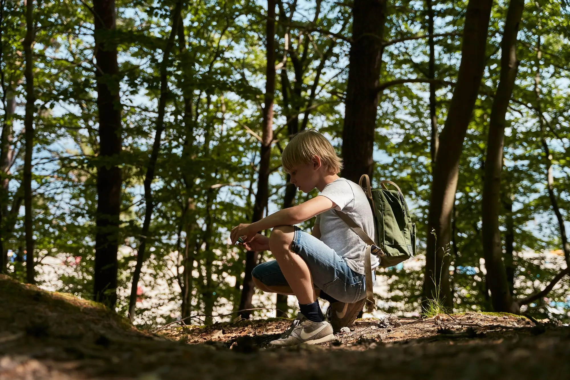 A boy kneels in the forest, surrounded by trees, wearing outdoor clothing and hiking boots.