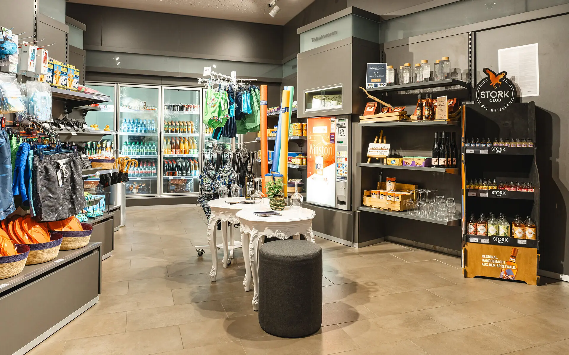 A small shop with cool drinks, bathing essentials and snacks. The room has grey walls and flooring and a warm light. 