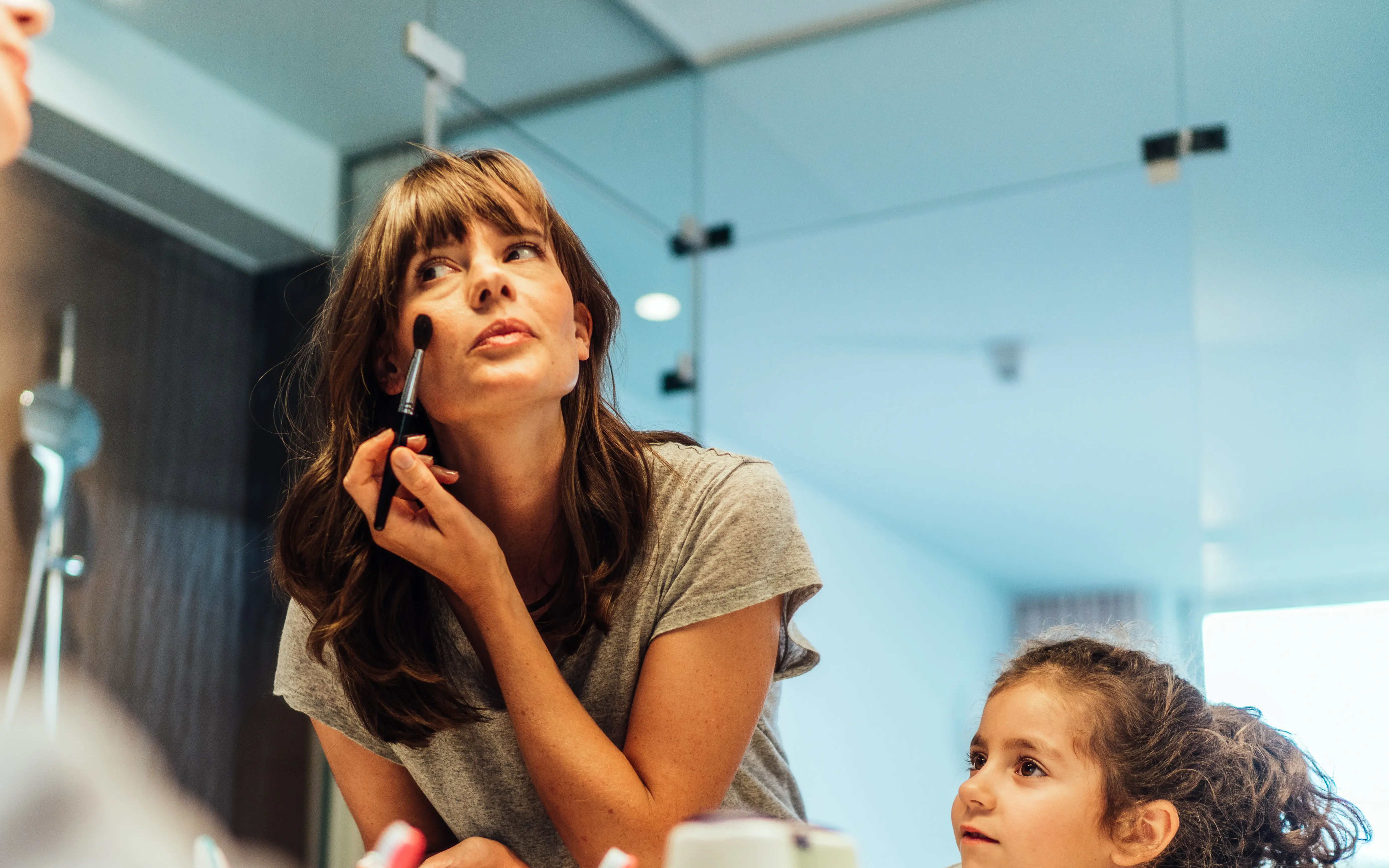 A mother applying makeup in front of a mirror while a child watches attentively.