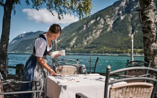 A woman in a dirndl sets a table by a lake.