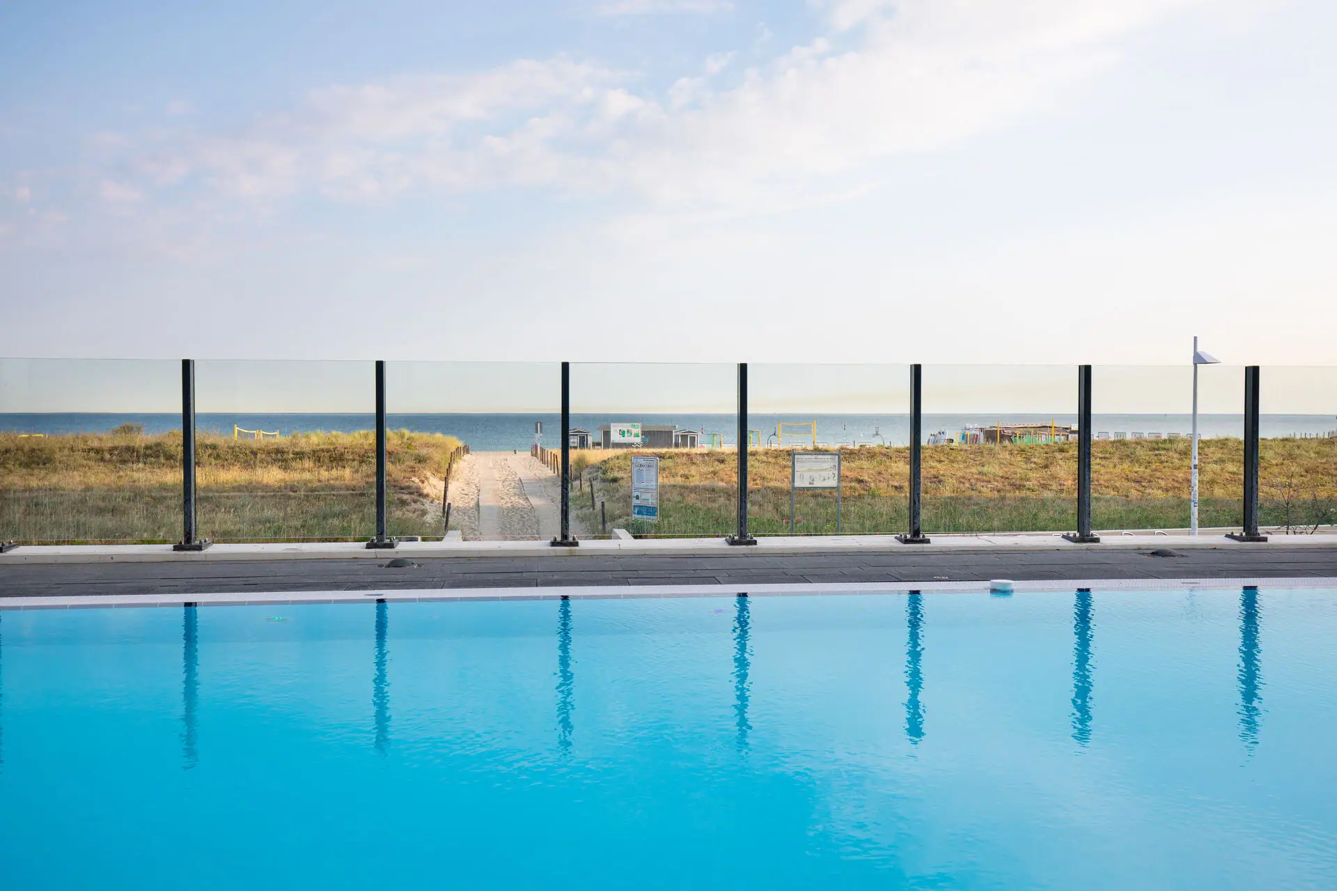 Outdoor pool at the aja Warnemünde A pool with a fence and lawn in the foreground, with a beach behind it.